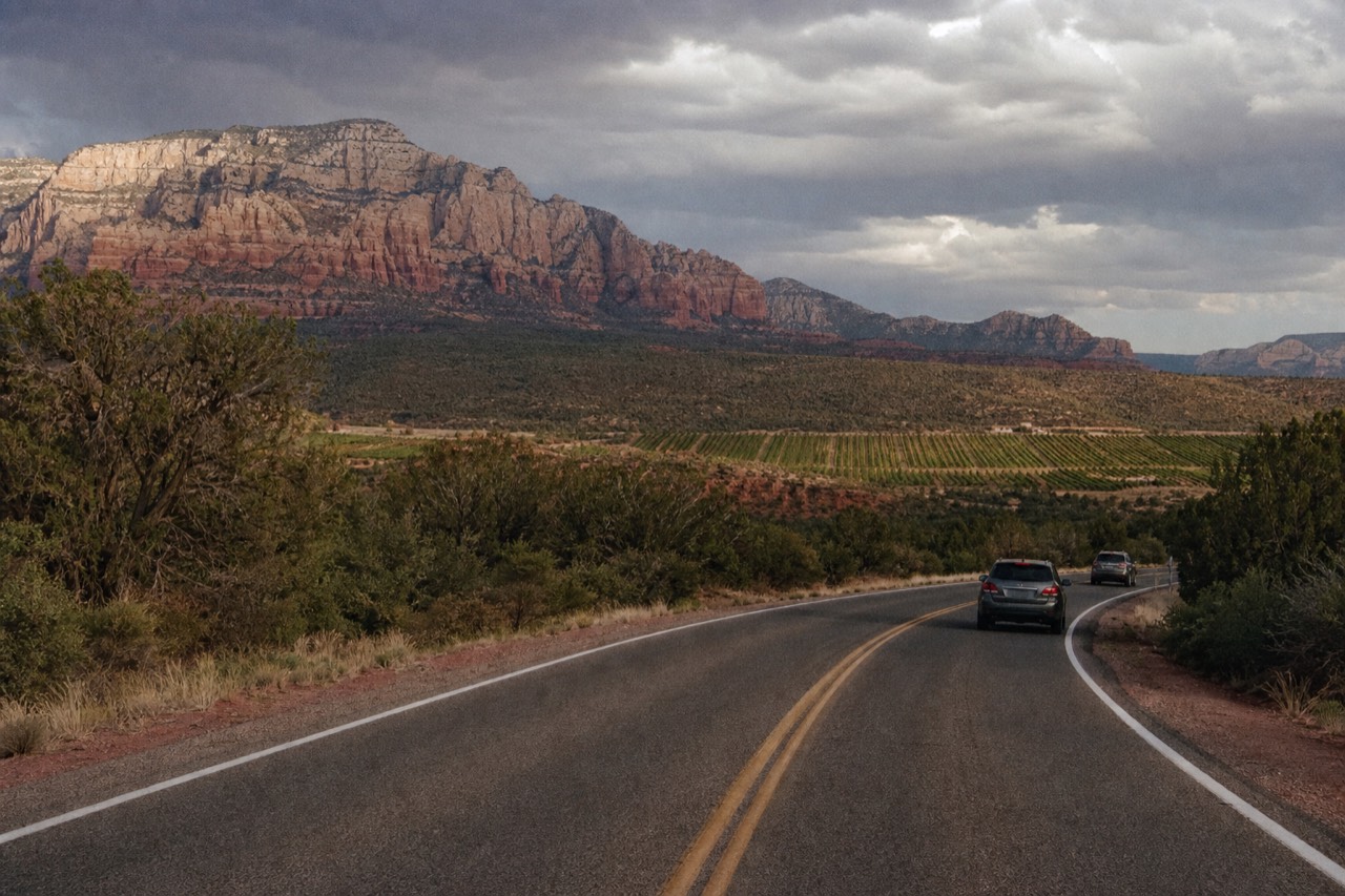A two-lane road curving through juniper scrub with red rock formations behind and vineyard rows visible in the valley ahead