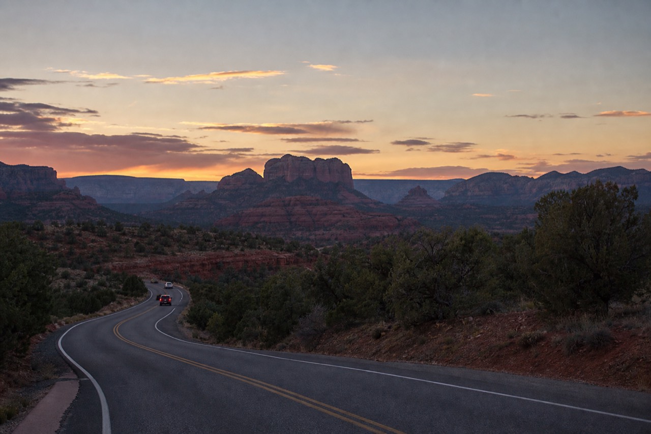Morning light across a Sedona valley with red rock formations layered into the distance and a two-lane road curving through juniper