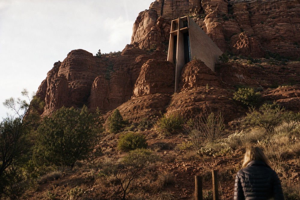 Small modernist chapel built into a red sandstone butte, seen from below in warm afternoon light with desert scrub around the base