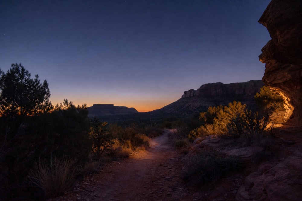 A narrow unpaved trail curving between scrub and red rock with no other people visible, early morning shadows across the ground