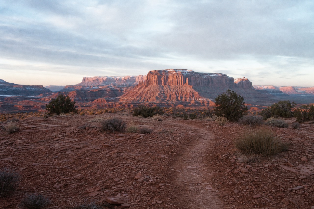 An empty red rock overlook at first light with a faint dirt path leading toward layered sandstone buttes in the distance