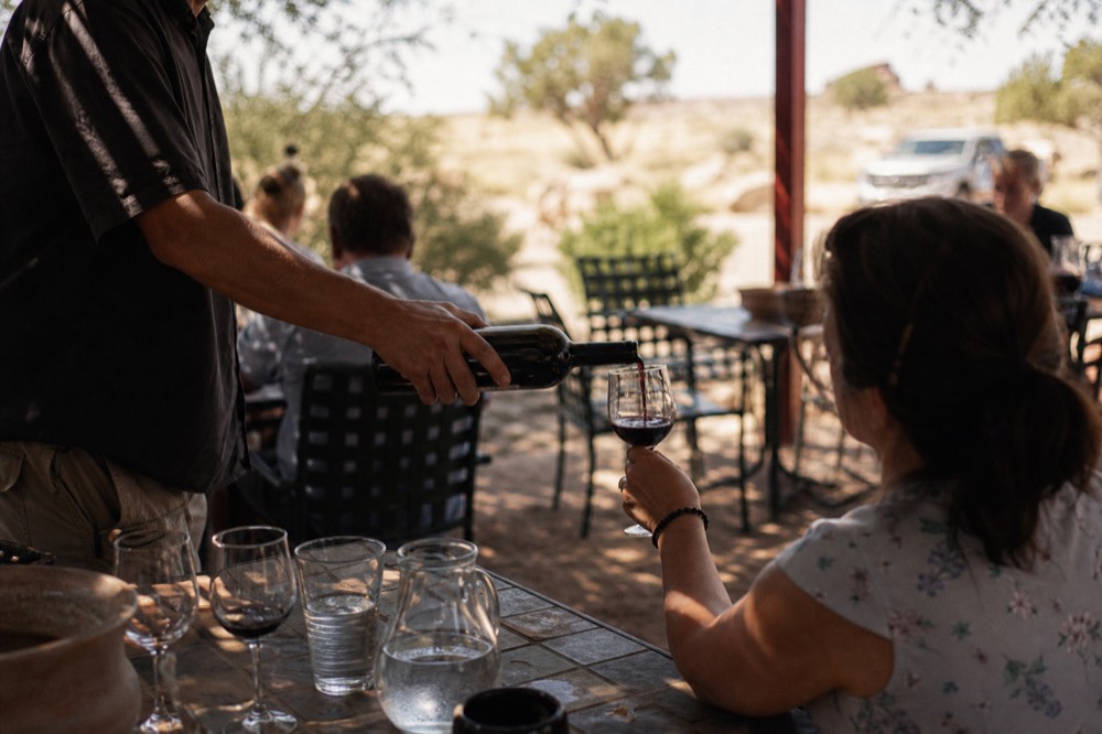 A guide pouring a small measure of red wine into a glass held by a seated guest at an outdoor table