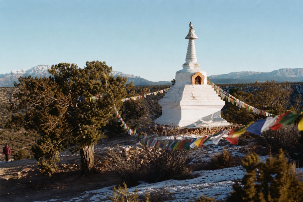 A whitewashed hillside stupa with prayer flags strung between junipers under a clear desert sky