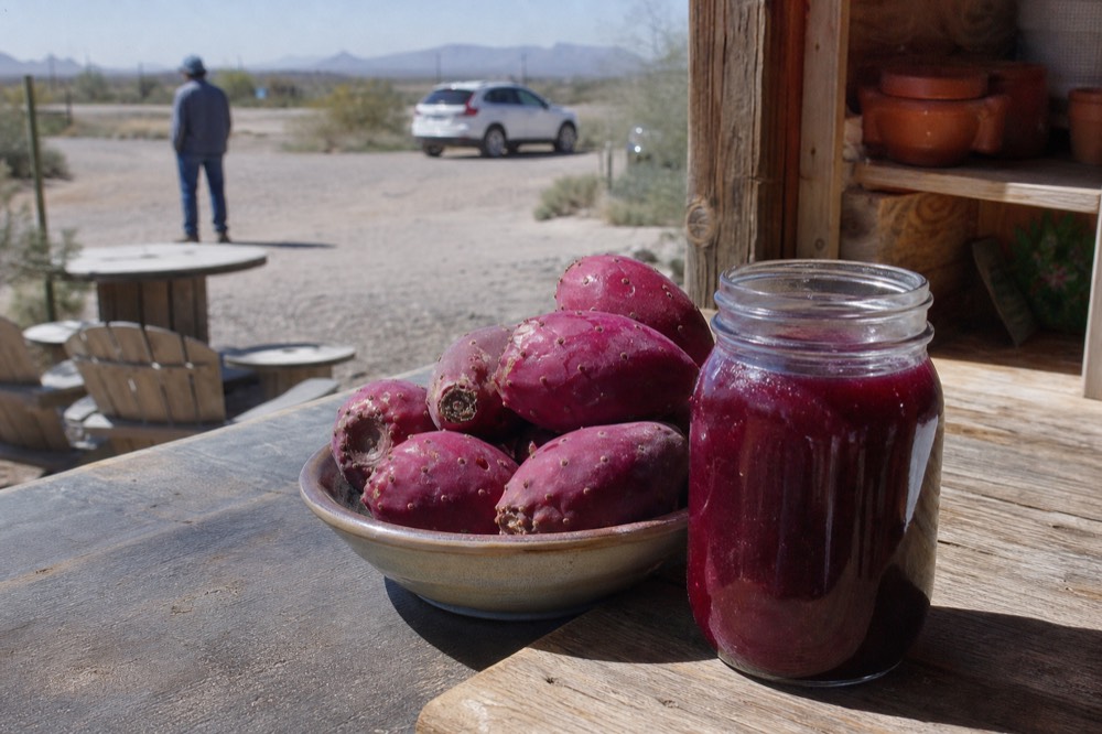 Deep magenta prickly pear fruit in a shallow bowl beside a jar of the same coloured syrup on a wooden counter