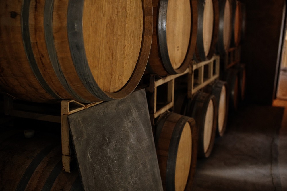 Stacked oak barrels in a dim cellar with a chalk-marked slate propped against the nearest row