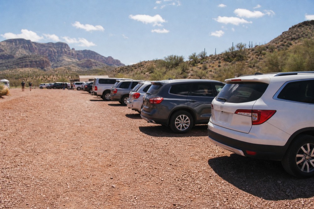 A full gravel trailhead parking area with vehicles parked along the shoulder of a red-dirt access road
