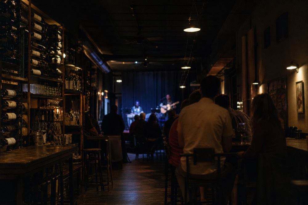 A narrow wine bar interior at night with bottles on shelves, a small corner stage and people at high tables
