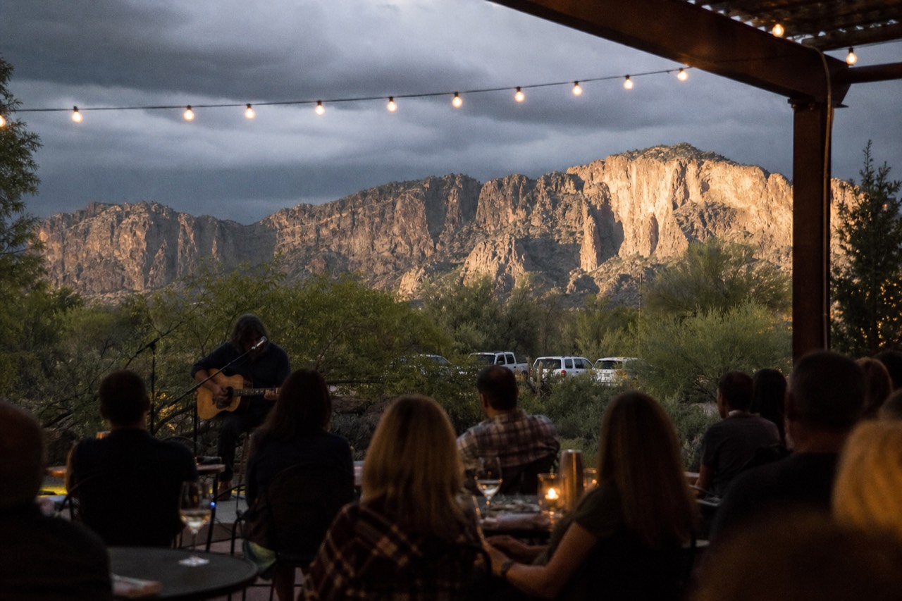A guitarist playing to a small seated crowd on a warm evening patio strung with bulb lights, wine glasses on the tables