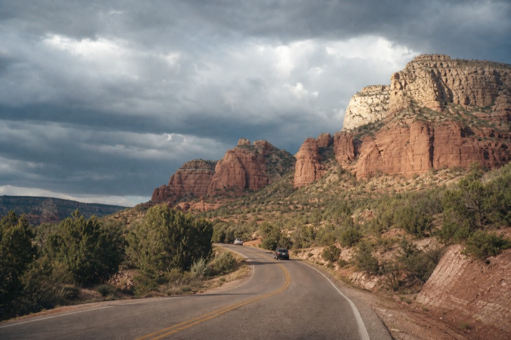 A two-lane desert highway curving between red rock buttes and juniper scrub under a wide afternoon sky
