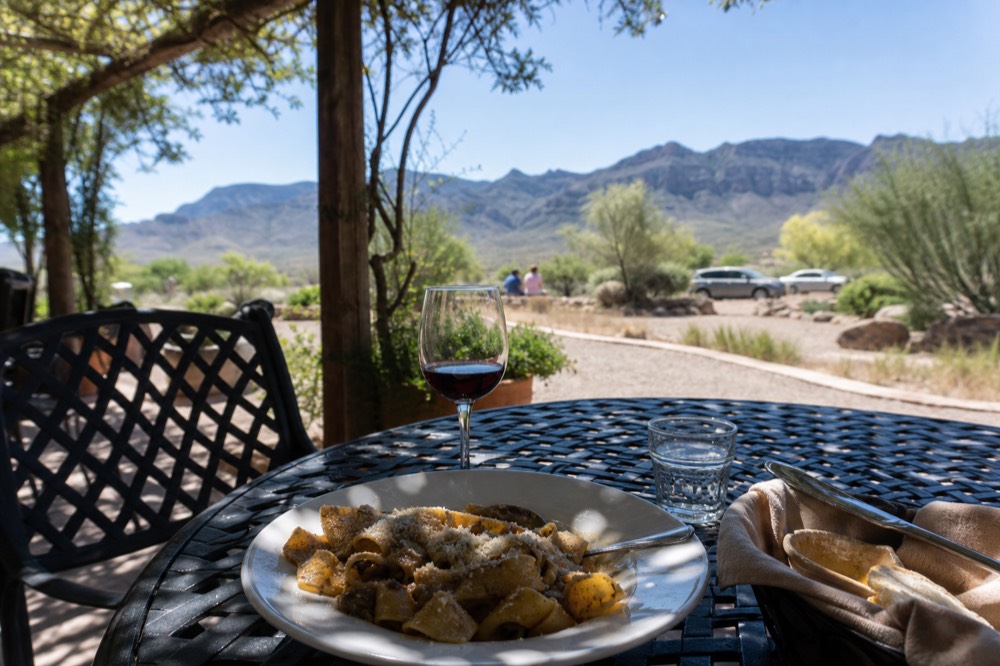 An outdoor patio table with a plate of pasta, a glass of red wine and a bread basket, dappled shade from a vine-covered pergola