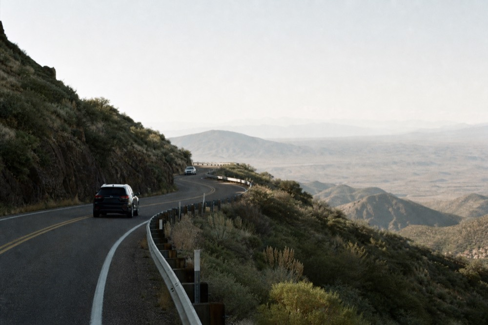 A narrow switchback road climbing a scrub-covered hillside with a guardrail and a wide valley falling away below