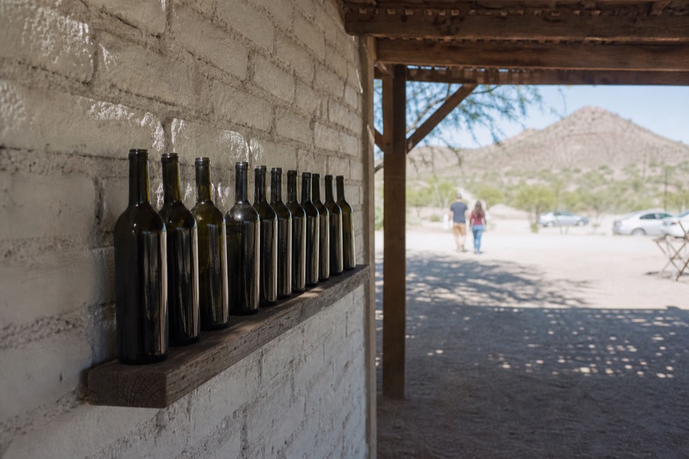A row of dark wine bottles standing on a rough wooden shelf against a whitewashed brick wall, no labels legible