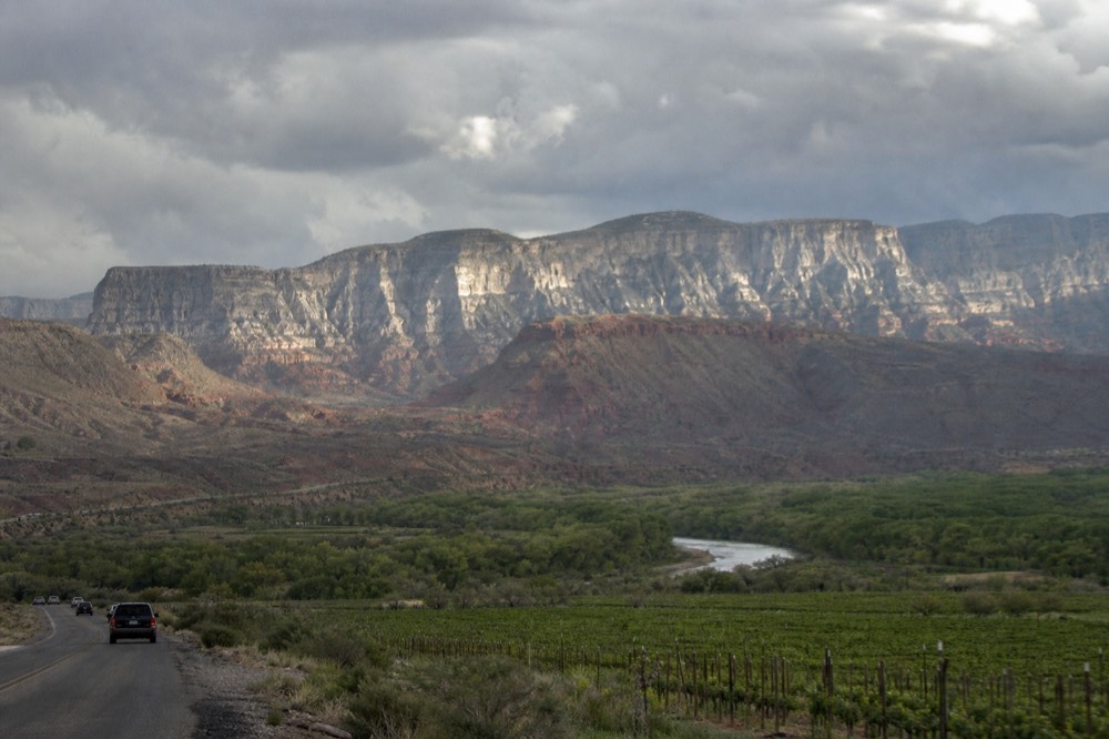 A wide view over a green river valley with vineyard blocks in the foreground and layered red and grey mesas behind