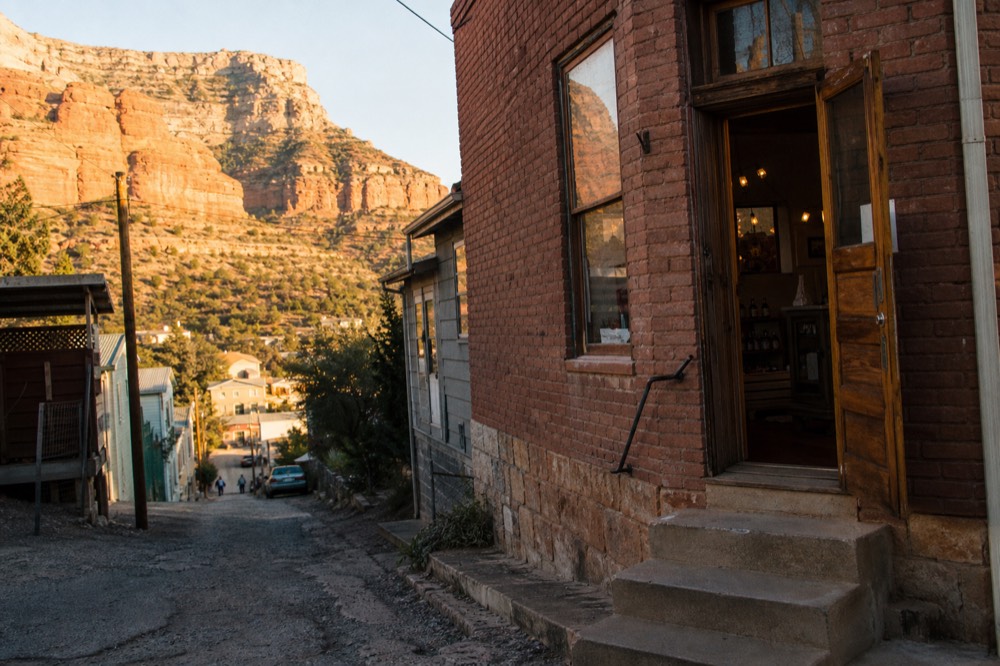 A weathered brick storefront on a steep narrow street in an old hillside town, with a wooden door open and warm light inside