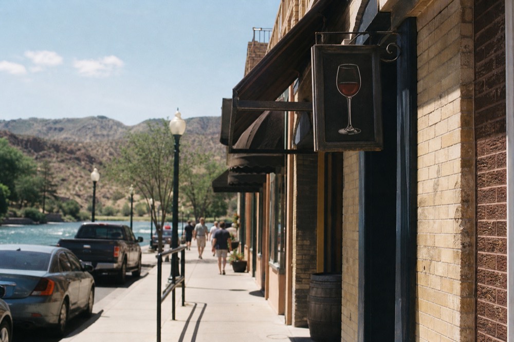 Wide sidewalk along a row of old brick storefronts with awnings, a hand-painted wine glass sign hanging above a doorway