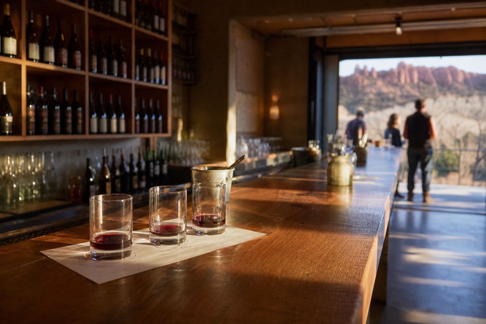 A narrow tasting room interior with a wooden bar, rows of bottles on shelves and three flight glasses on a paper tasting mat