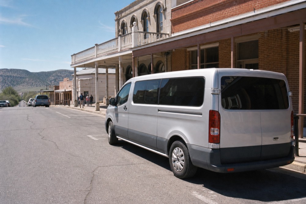 A small silver passenger van parked at an angled kerbside space on a historic main street lined with low brick buildings