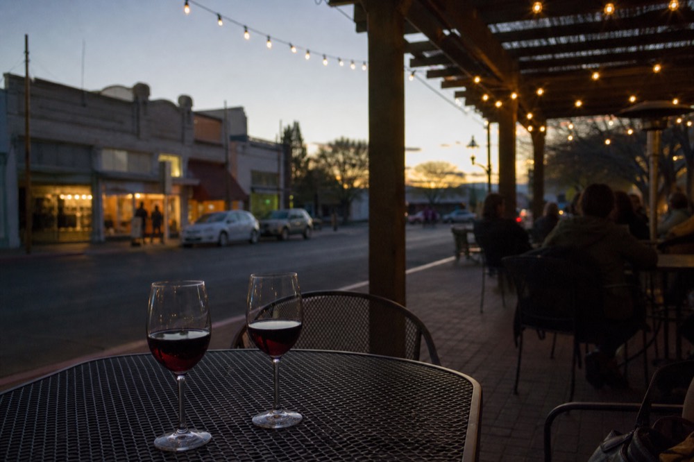 Evening on a small-town street with warm window light from storefronts, string lights overhead and glasses of red wine on an outdoor table