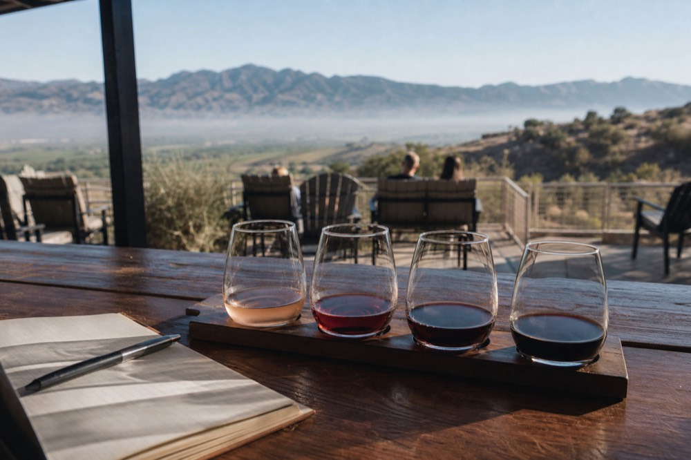 A flight of four small wine pours lined up on a wooden paddle with a notebook and pen beside it on a bar