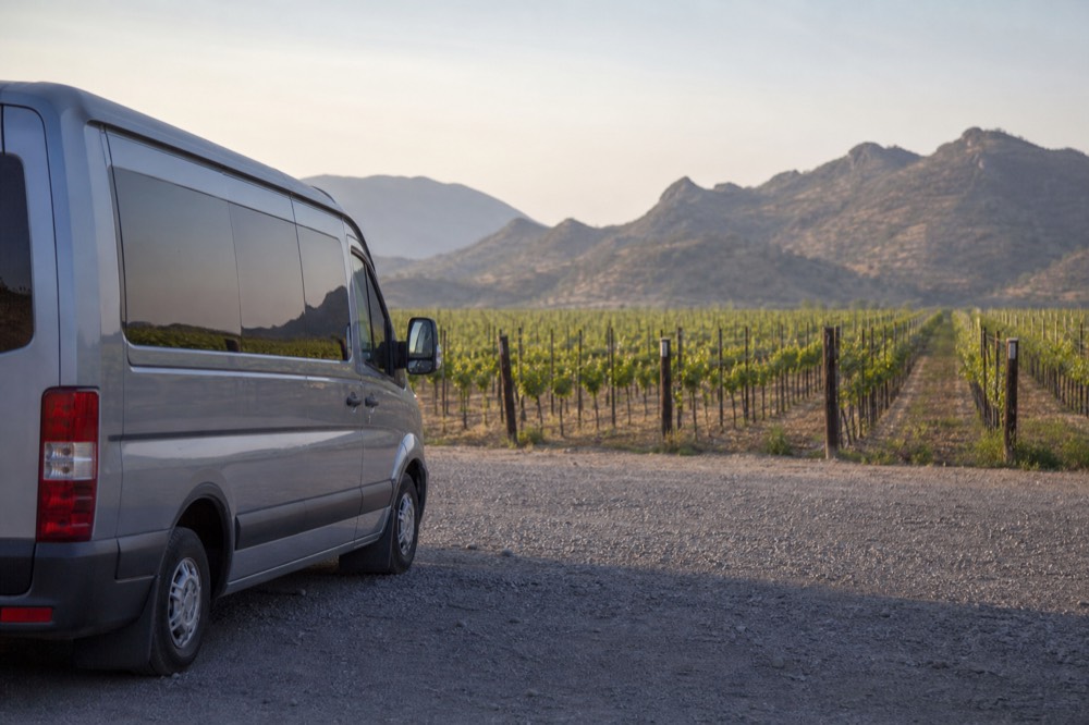 A small tour van parked on gravel beside a vineyard fence with green rows stretching to a low ridge