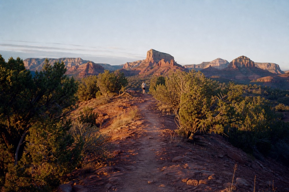 Narrow dirt path climbing a low red rock knoll at sunrise with junipers on both sides and layered buttes behind