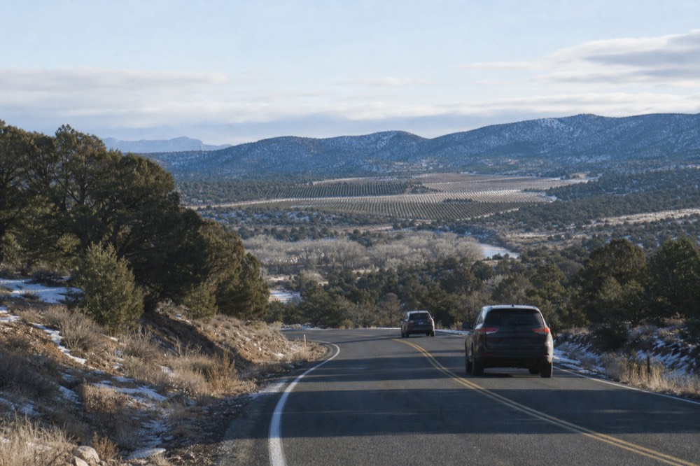 Two-lane road curving down through juniper hills toward a green creek corridor with vineyard rows visible in the distance