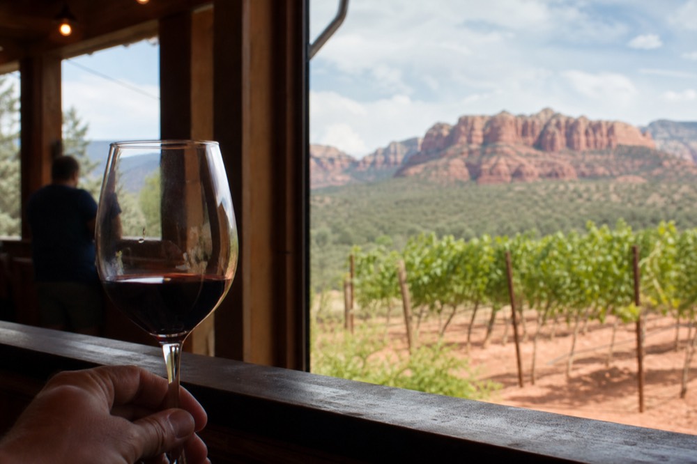 Hand holding a stemmed glass of red wine on a wooden rail with sunlit vineyard rows and red cliffs beyond