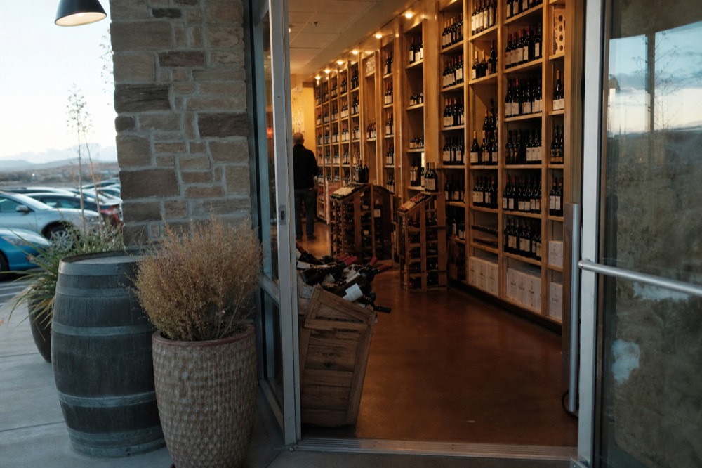 Floor-to-ceiling wooden wine shelves packed with bottles in a small retail wine shop, warm overhead lighting, no signage visible