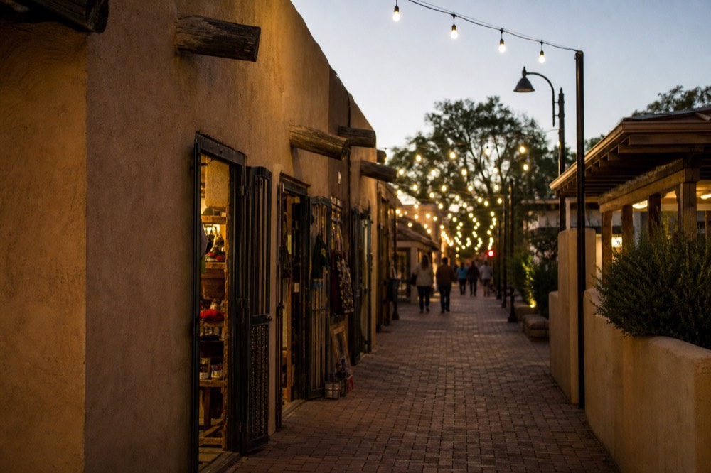 A narrow southwestern shopping arcade at dusk with adobe walls, string lights overhead and open shop doorways