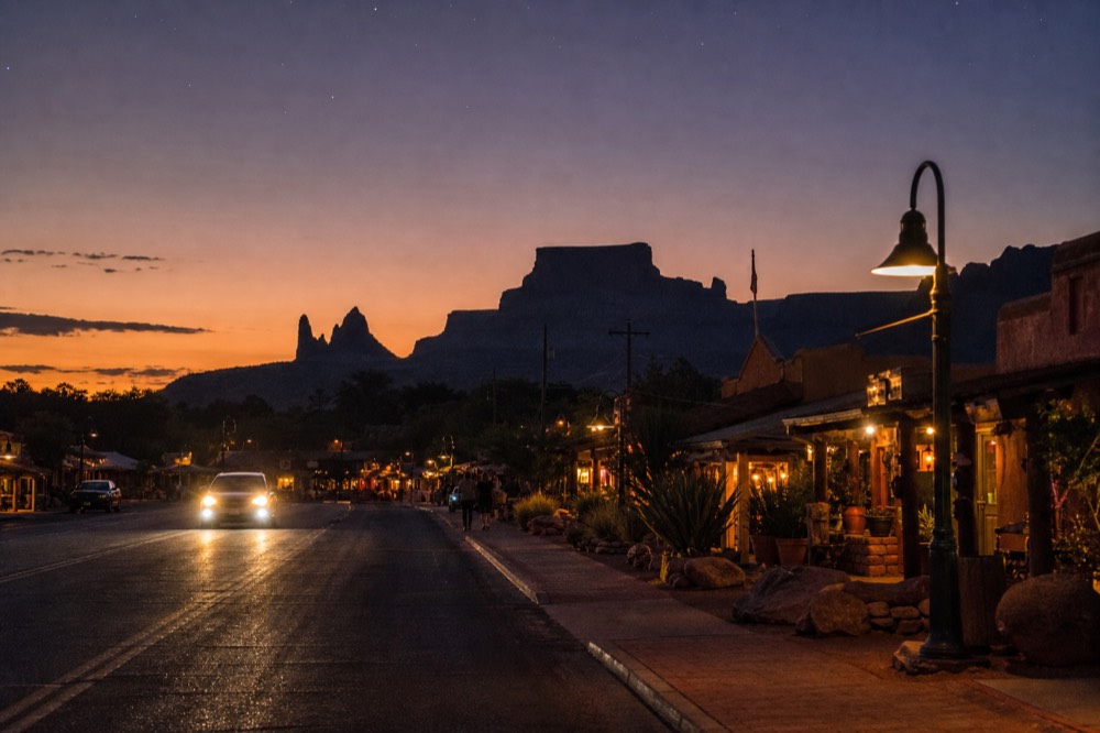 Evening view along a desert highway strip with low buildings, warm shopfront lights and red rock silhouettes against a fading orange sky