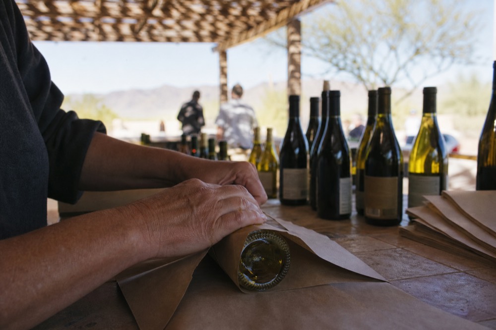 Close-up of hands wrapping a wine bottle in paper on a shop counter with more bottles blurred in the background