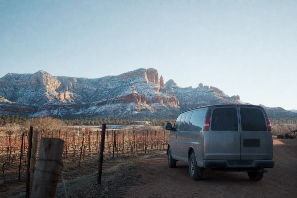 Passenger van parked on a dirt shoulder beside a vineyard fence with red cliffs rising in the distance