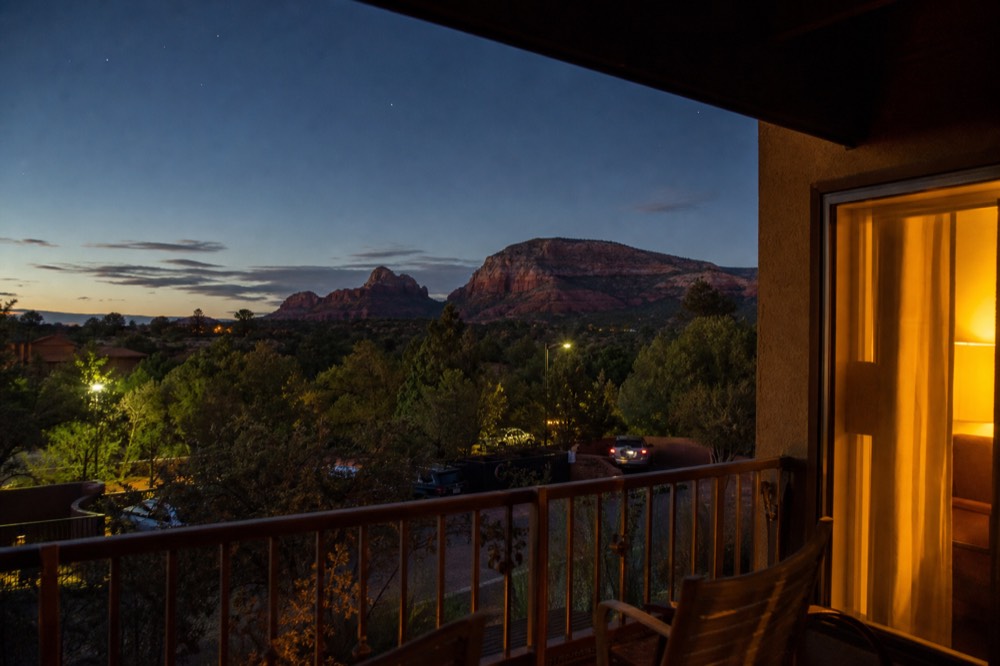 Evening view from a hotel balcony over juniper and red sandstone buttes with warm interior light spilling onto the rail