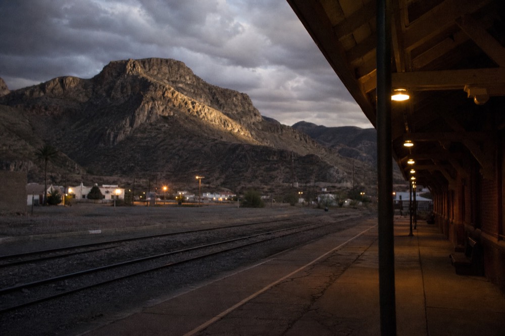 Empty small-town rail depot platform at dusk with warm lamps along the boarding edge and desert hills behind