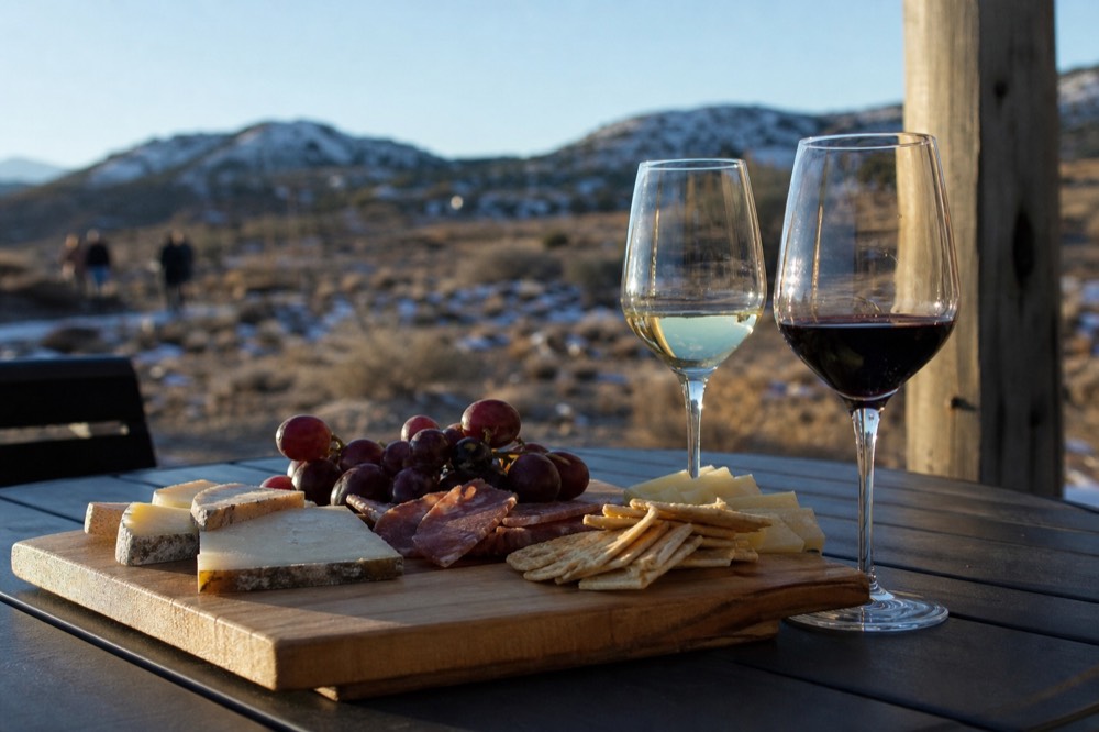 Wooden board with sliced hard cheeses, cured meat, grapes and crackers on a table beside two wine glasses