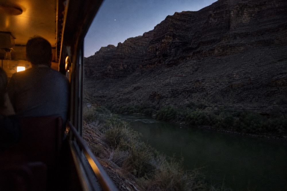 View from a moving train of steep layered canyon walls above a narrow green river with no road visible
