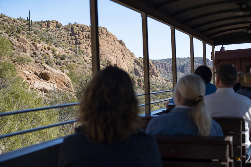 Open-air rail viewing car with a metal rail and bench seats, passengers' silhouettes against sunlit canyon rock