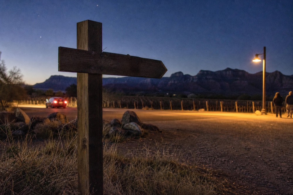 A wooden trail signpost at a gravel pull-off pointing along a rural road, dry grass in the foreground and trellised vines in the middle distance