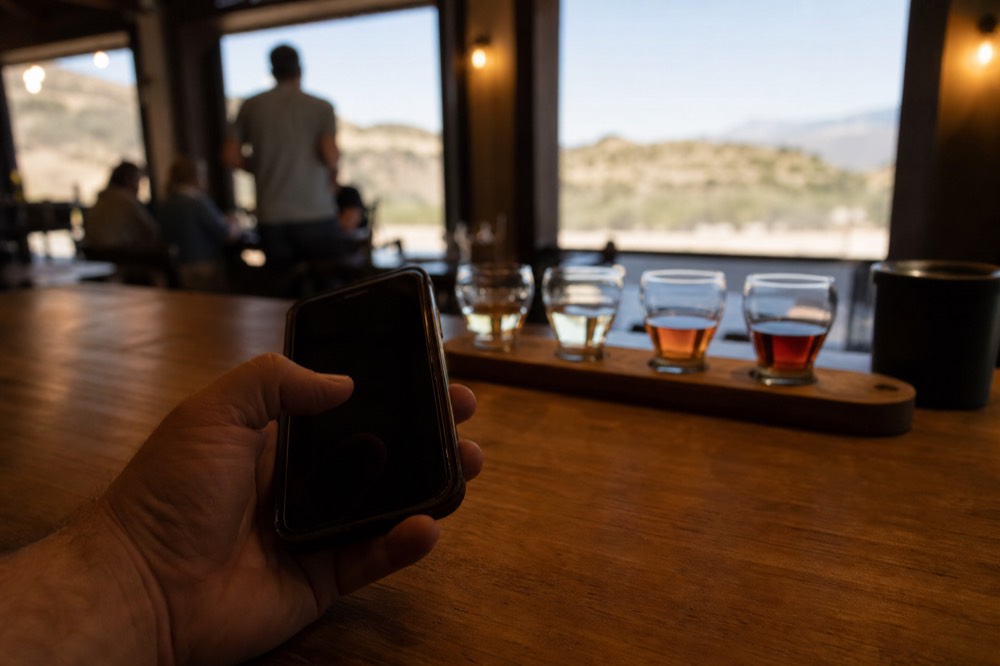 A hand holding a phone at a wooden tasting bar, a tasting flight of four glasses out of focus behind it in warm indoor light