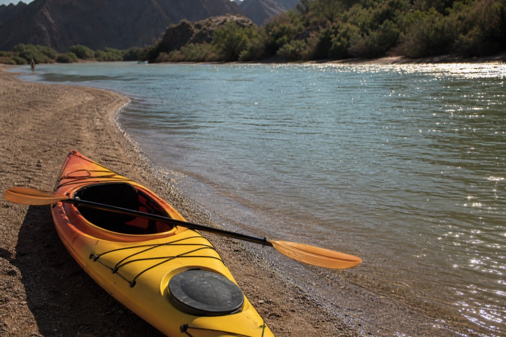 An empty kayak resting on a sandy riverbank in warm evening light with a paddle laid across the cockpit