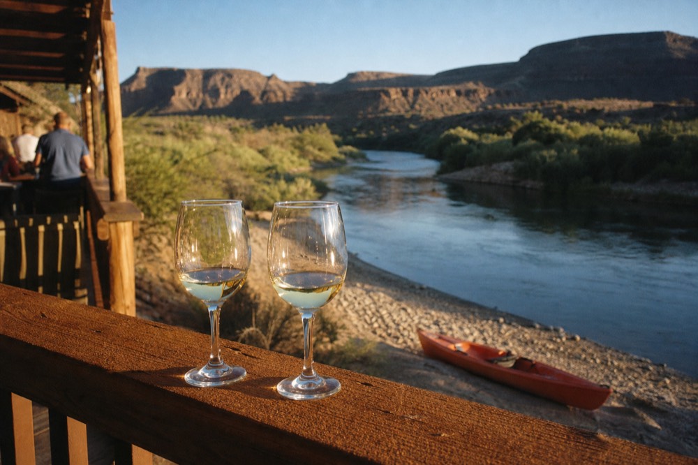 Two wine glasses on a wooden rail overlooking a river valley at golden hour with a kayak drawn up on the bank below