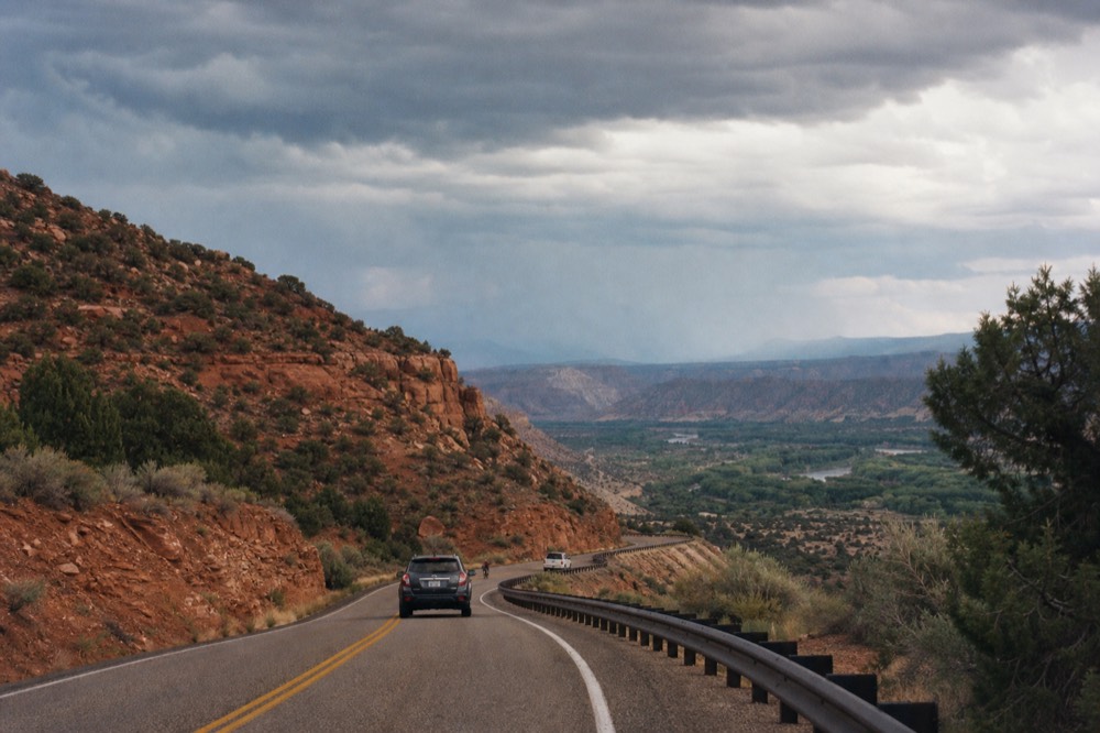 A two-lane road curving down from red rock country toward a green river valley, guardrail on one side and juniper scrub on the other