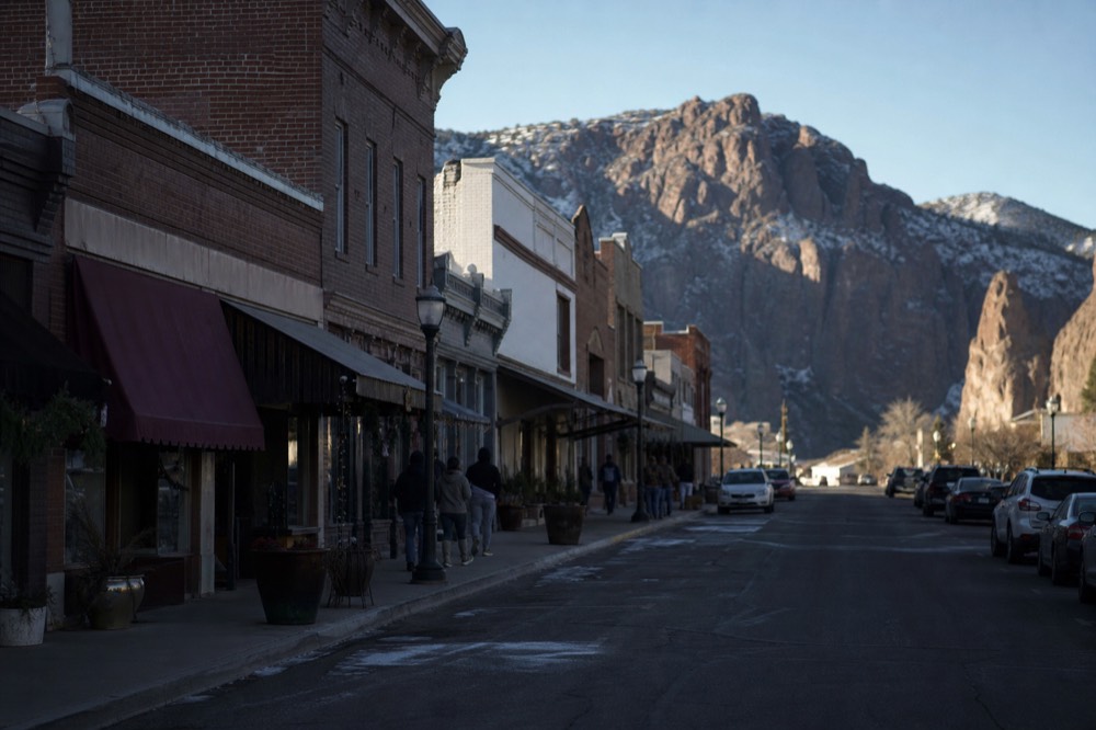 A historic small-town main street of low brick storefronts with awnings, a few people walking between doorways in late afternoon shade
