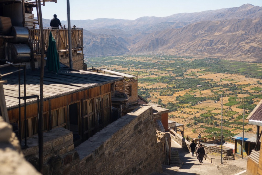A steep hillside town of stacked wooden and brick buildings clinging to a mountainside, with a wide valley of green and tan visible far below